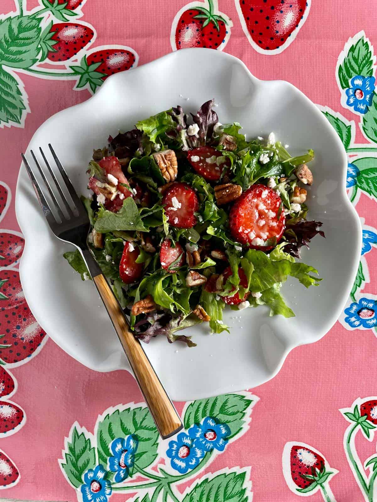 A white bowl of strawberry salad, pecans, and feta with a wooden handled fork.