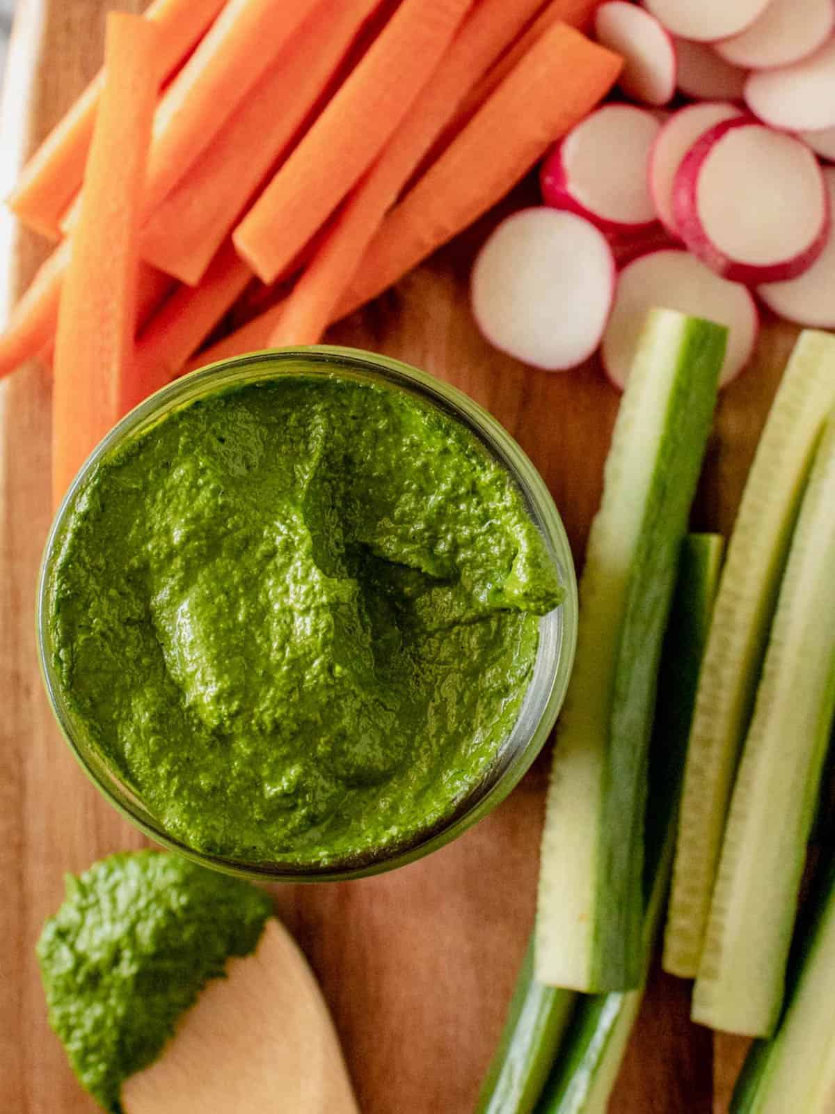 Green tahini sauce in a jar with carrots, radishes, and cucumbers.
