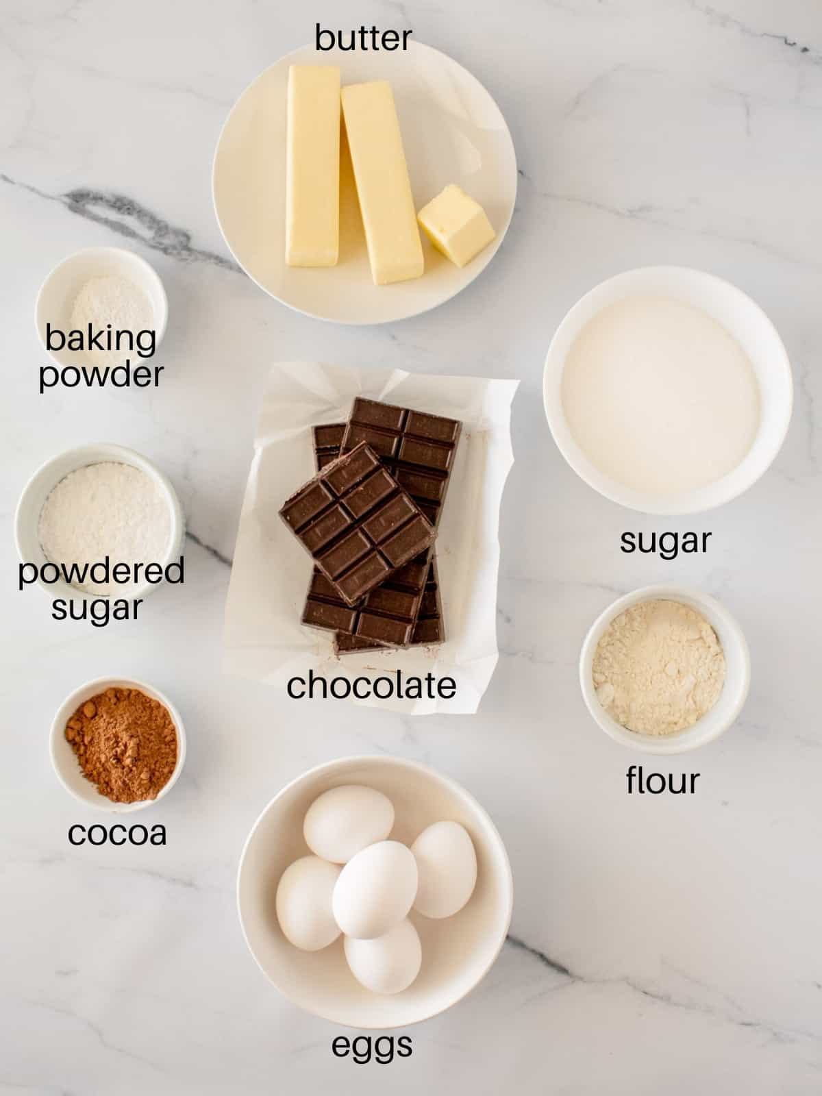 Ingredients in small bowls including butter, sugar, bar chocolate, flour, cocoa, and eggs.
