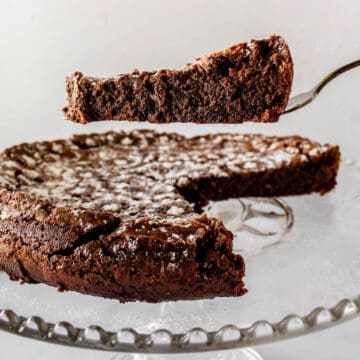 Chocolate cake on pedestal with slice on server hovering above revealing fudgy texture.