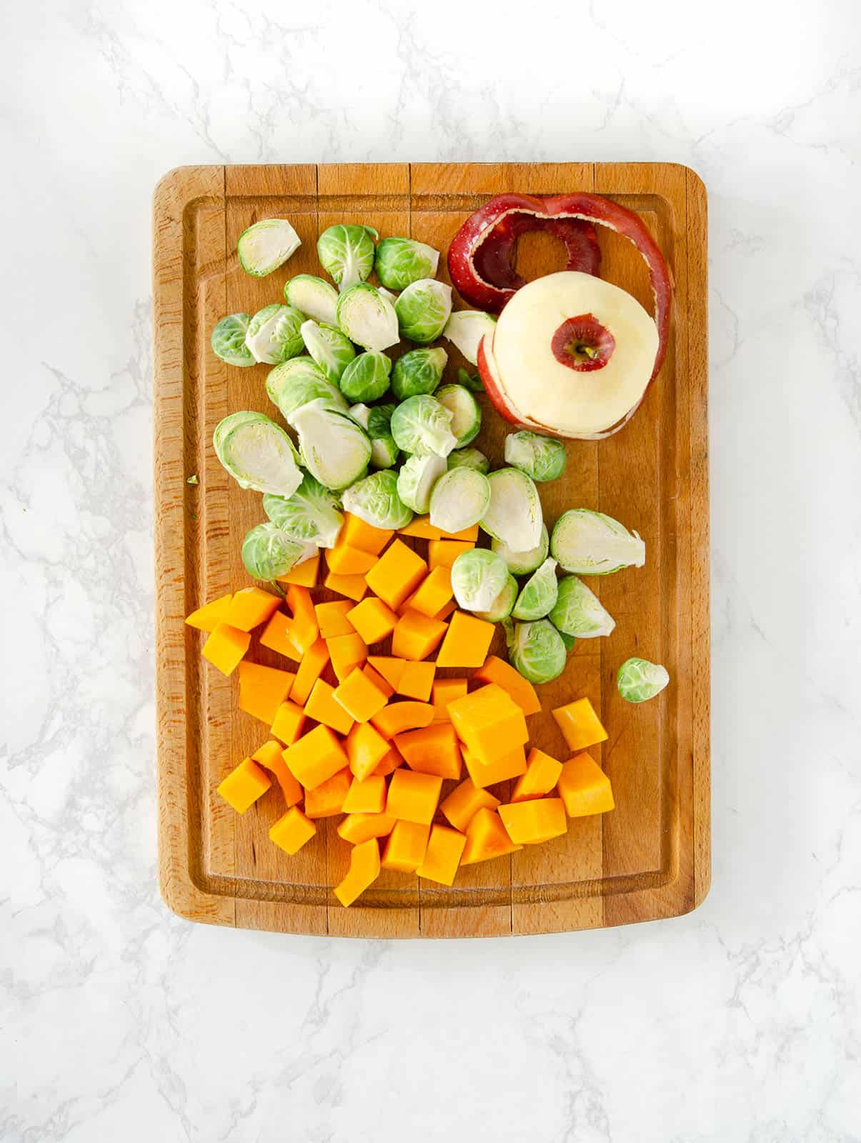Chopped butternut squash, sliced Brussels sprouts, and peeled apple on cutting board.