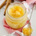 A jar of homemade applesauce with cinnamon sticks and an apple in the background.