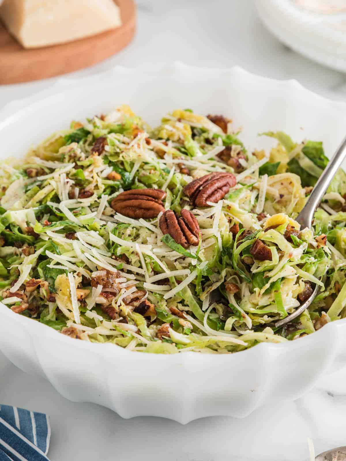 Shaved Brussels sprouts salad with parmesan and pecans in white bowl with spoon.
