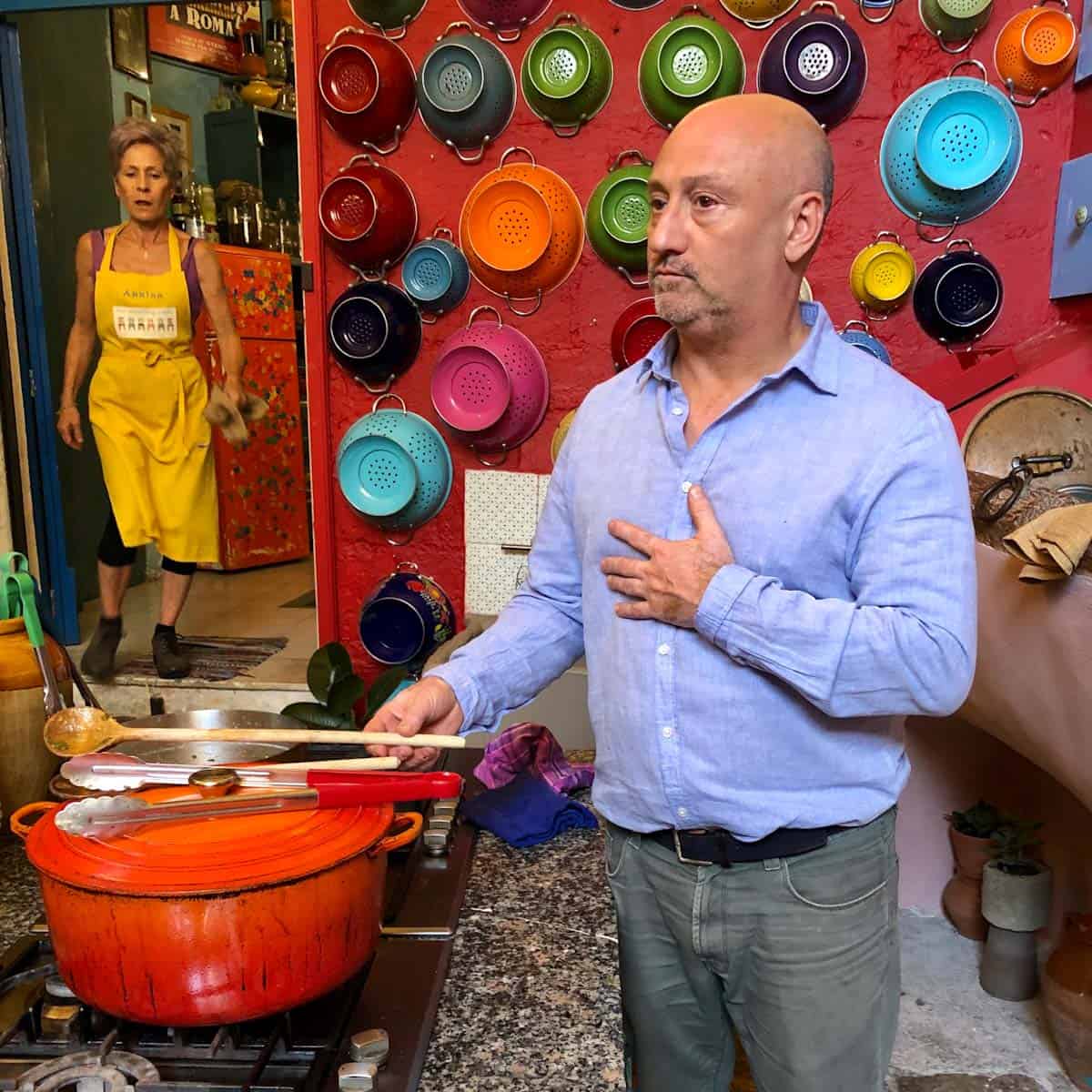 Silvestro at the stove in the colorful Awaiting Table Cooking School kitchen with colanders on wall.