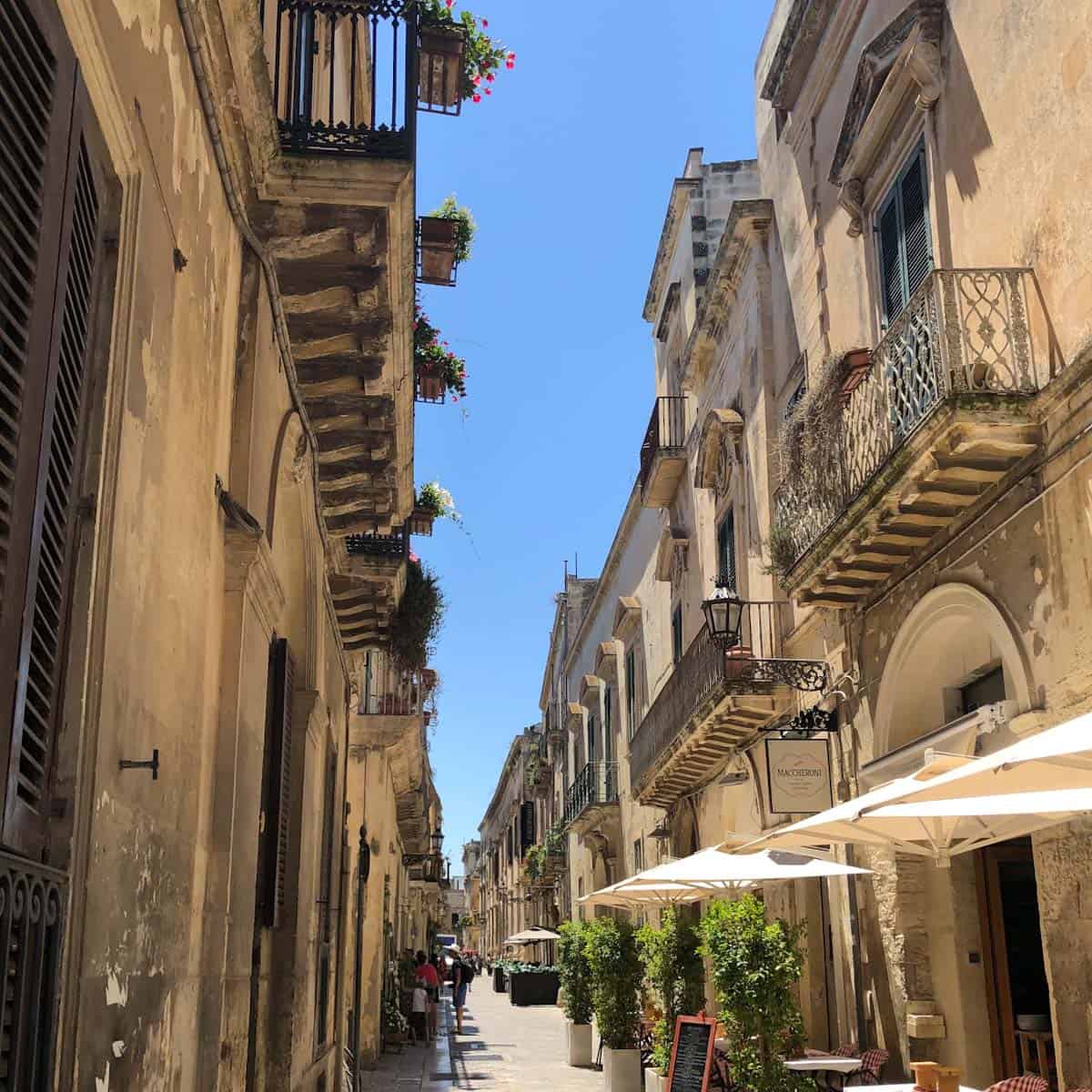 A narrow walking street in Lecce, Italy with Baroque carved buildings and outdoor restaurant tables.
