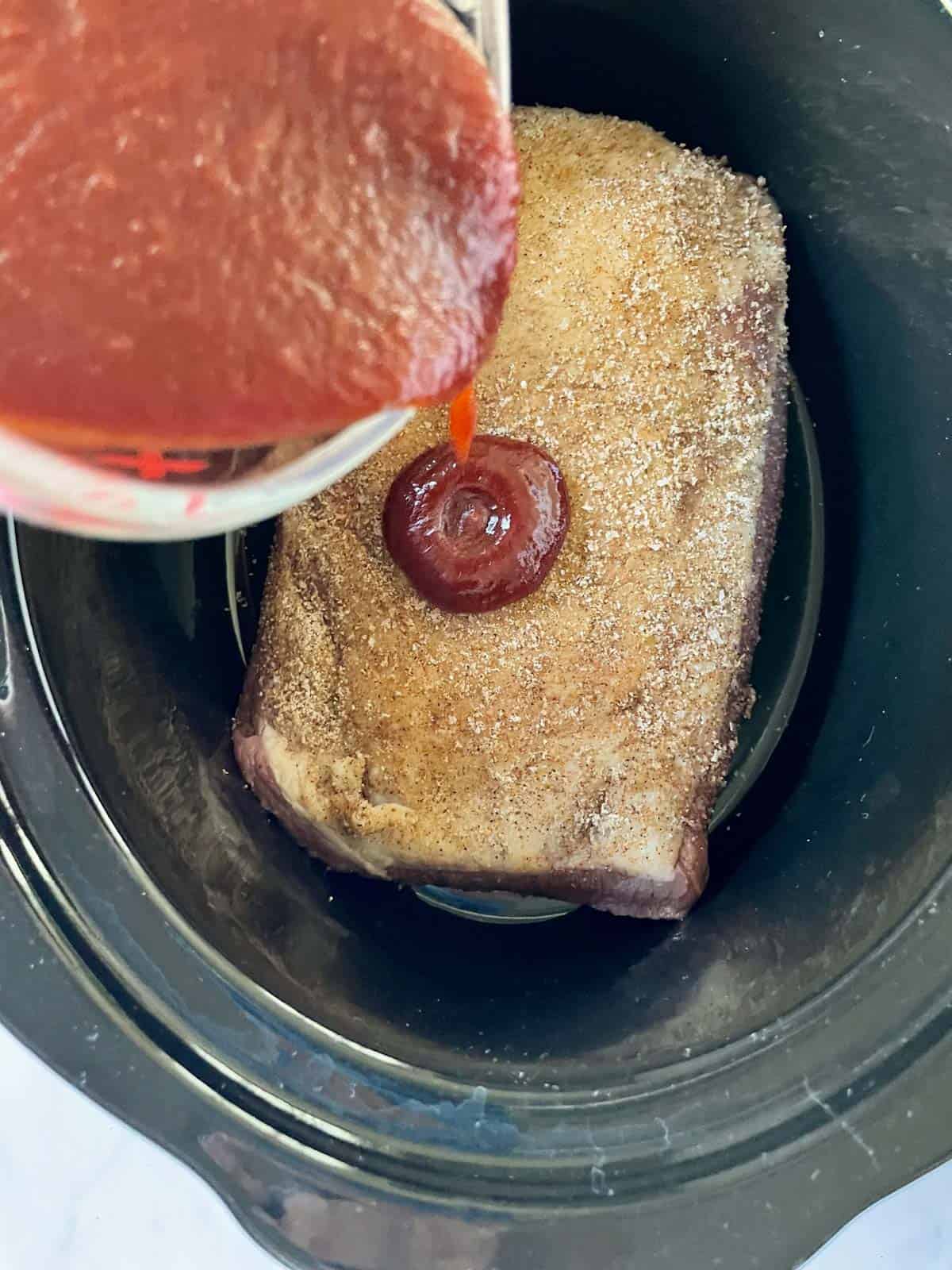 Pouring BBQ sauce over a spice rubbed brisket in a slow cooker.