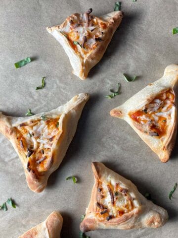 Baked hamantaschen pizzas on parchment paper topped with basil.