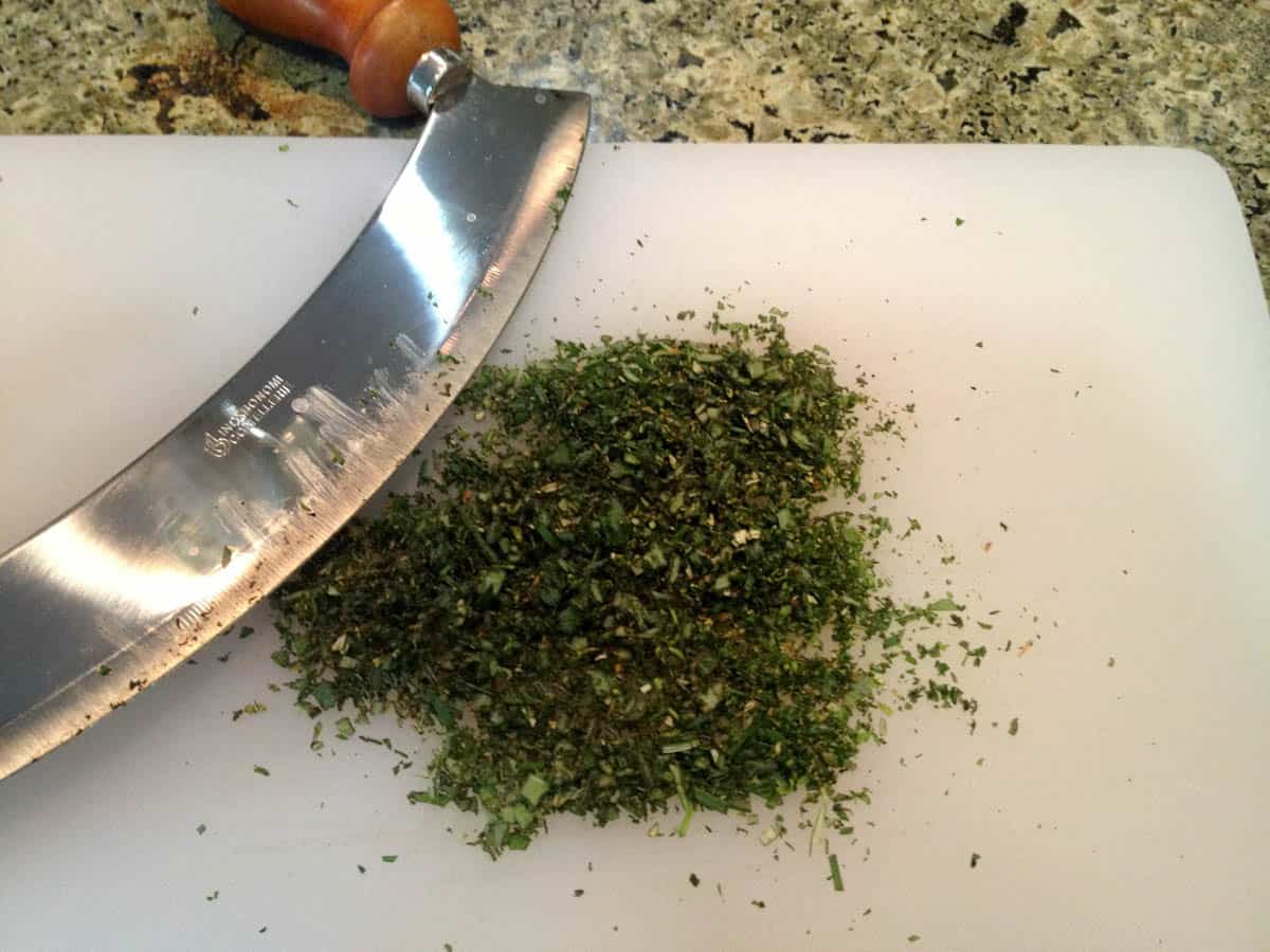 Finely chopped mixed herbs on a cutting board with a mezzaluna.