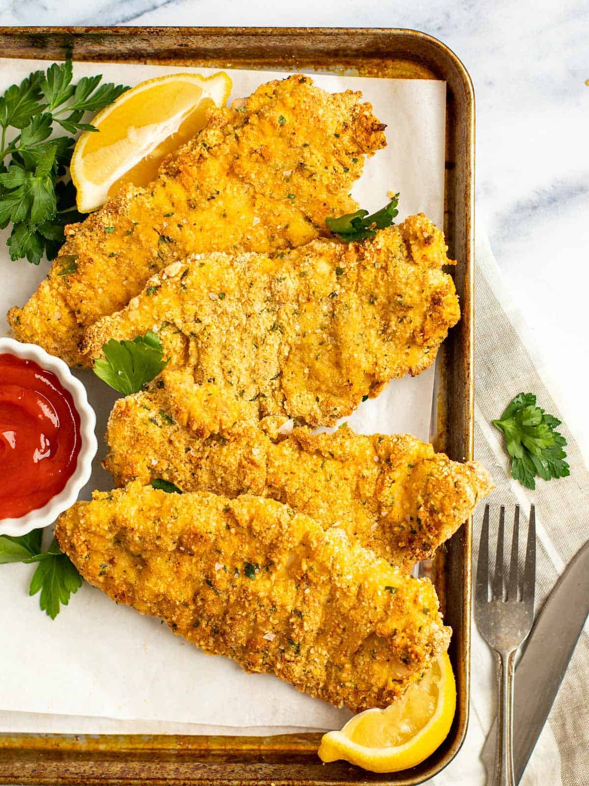Crispy chicken schnitzel on a baking tray with parsley, lemon, and a dipping sauce.