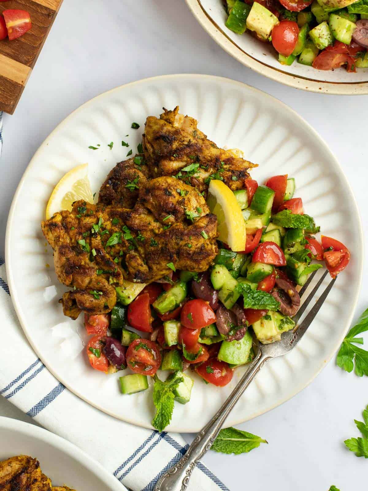 Mediterranean grilled chicken thighs on a plate with cucumber and tomato salad.