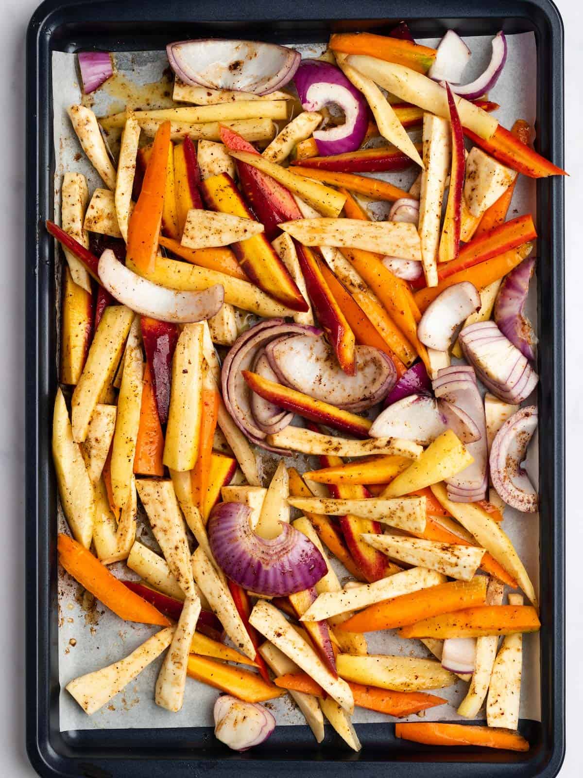 Carrots, onions, and parsnips on a baking sheet with parchment tossed in seasonings.