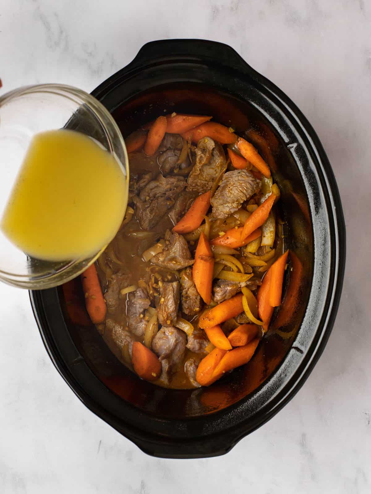 Pouring chicken broth into a slow cooker with lamb, carrots, and onions.