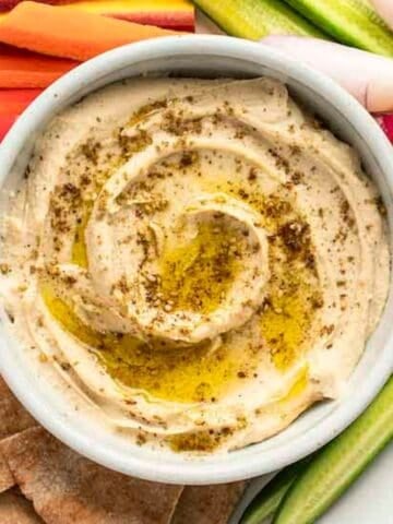 Hummus in a white bowl with sliced vegetables and pita surrounding it.