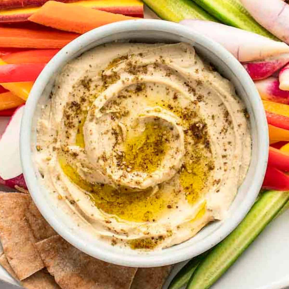 Hummus in a white bowl with sliced vegetables and pita surrounding it.
