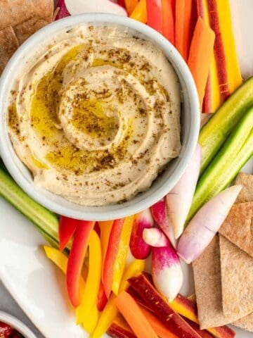 Hummus in a white bowl with carrots, celery, cucumbers, peppers, and shallots around the bowl.